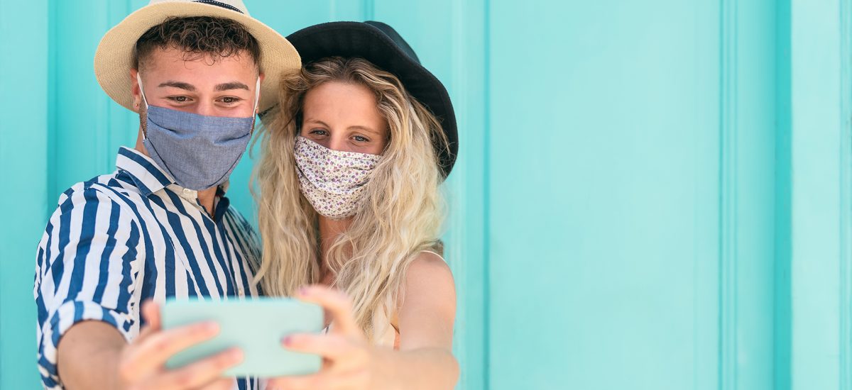 Young couple wearing face mask taking selfie with mobile smartphone on vacation - People having fun traveling again during corona virus outbreak - Love relationship and technology concept
